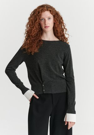 Femme aux cheveux roux bouclés portant un pull gris foncé avec des poignets blancs et un pantalon noir, debout, une main sur la hanche.