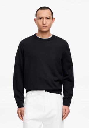 Mann mit kurzen Haaren, der einen schwarzen Langarmpullover mit rundem Ausschnitt und weiße Hosen trägt, vor einem schlichten weißen Hintergrund steht.