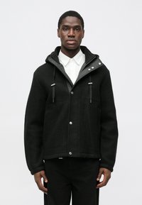 Sort uldjakke med hætte, sølvfarvede trykknapper og snoretræk. Har to lommer foran og et glat, tekstureret stof.