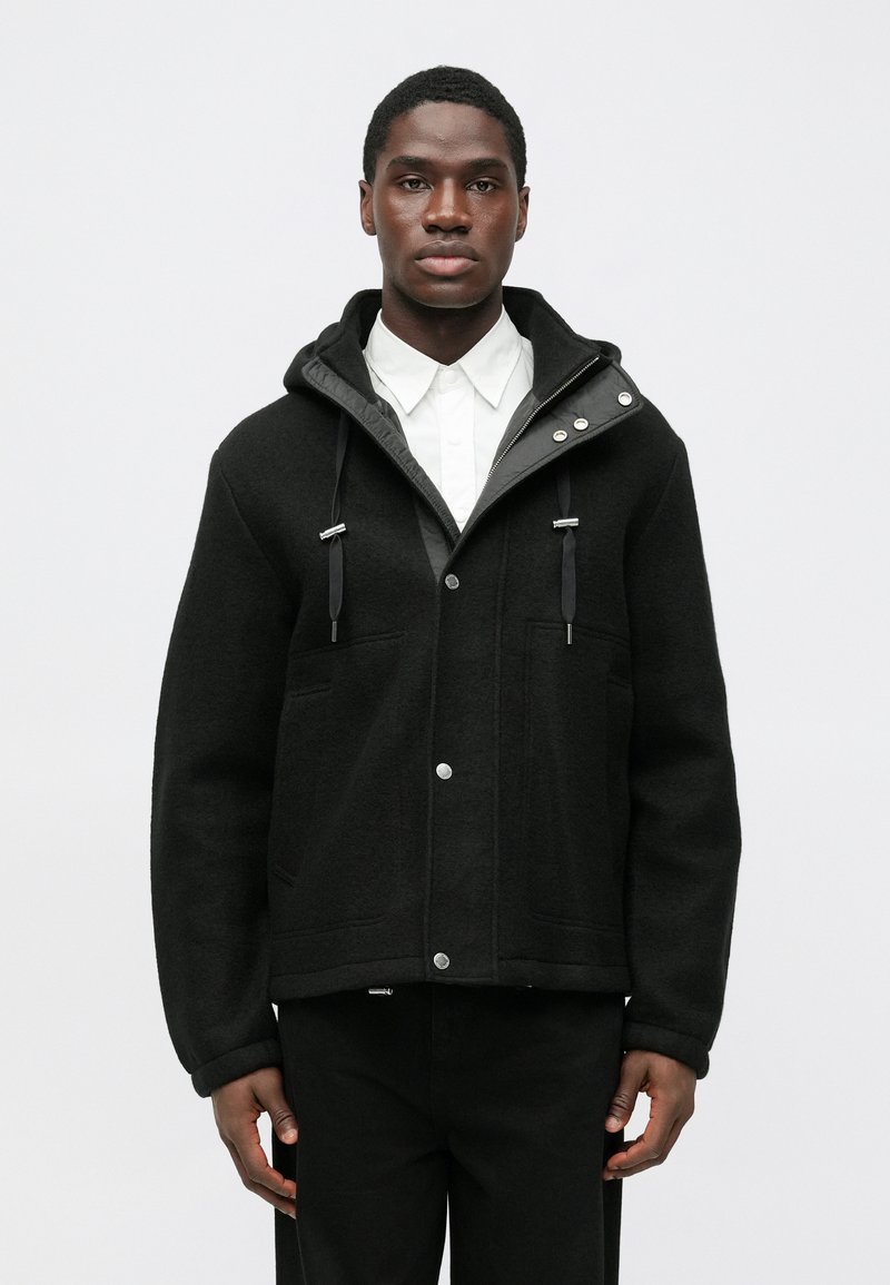 Sort uldjakke med hætte, sølvfarvede trykknapper og snoretræk. Har to lommer foran og et glat, tekstureret stof.