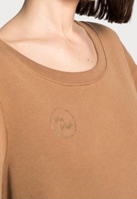 Brun sweatshirt med en rundad nacke och korta ärmar. Har en cirkulär logotyp i en ljusare nyans framme vid nacken.