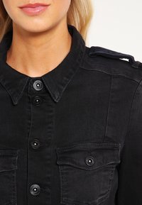 Svart denimskjorta med knäppning, två bröstfickor och axelremmar; textil med subtila sömnadsdetaljer.