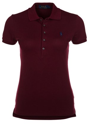 Žena, která nosí krátký burgundský polo tričko, černé přiléhavé kalhoty a černé kotníkové boty, stojící s jednou nohou vpřed.