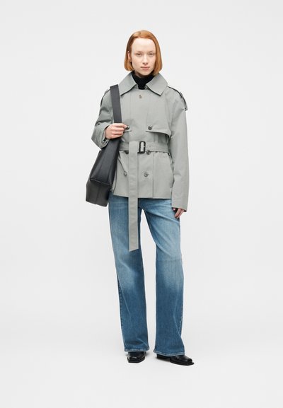 Νεαρό άτομο που φοράει γκρι ανοιχτόχρωμο παλτό τύπου trench με ζώνη, μπλε τζιν φαρδιάς γραμμής, μαύρα παπούτσια, μαύρη τσάντα ώμου, στέκεται μπροστά σε λευκό φόντο.