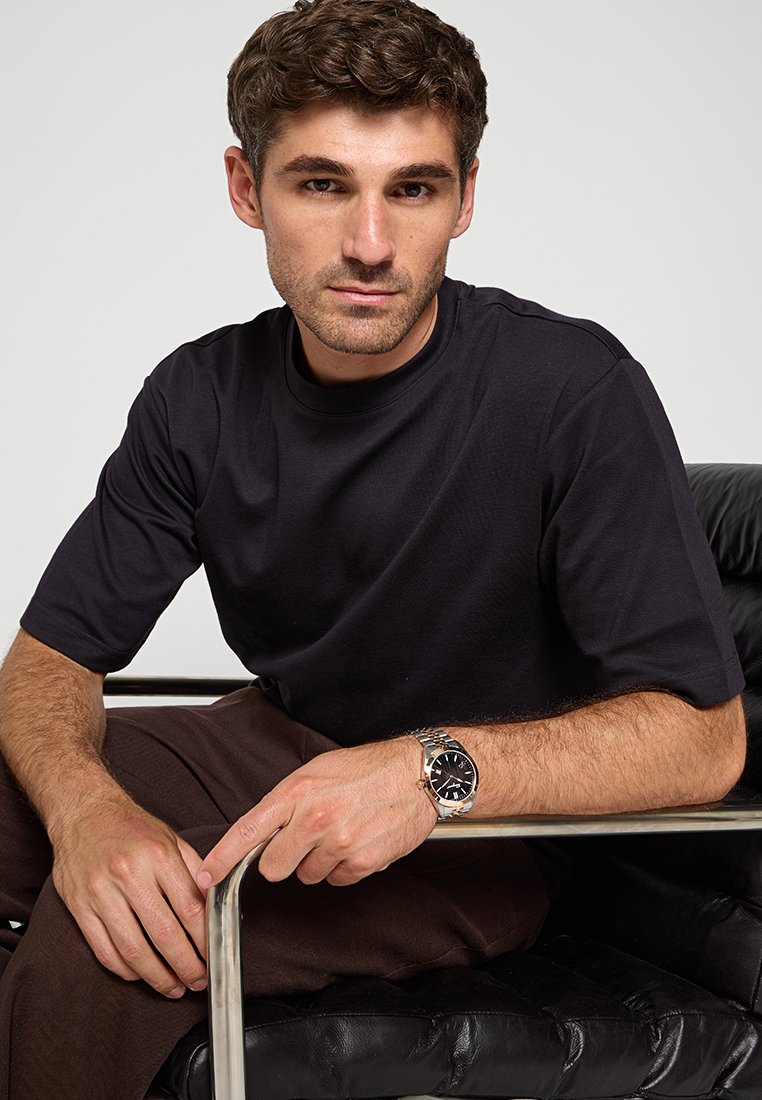 Camiseta negra de manga corta, tela suave. Pantalones marrones. Reloj de pulsera plateado con una esfera negra. Modelo sentado en una silla de cuero negra.