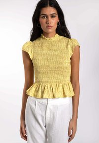 Top peplum amarillo con volante cortos y cuello alto. Tela texturizada con un patrón sutil, combinada con pantalones blancos de tiro alto.