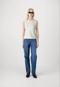 Γυναίκα με αμάνικο λευκό μπλουζάκι, ίσιους μπλε τζιν, μαύρα παπούτσια και μαύρα γυαλιά ηλίου, που στέκεται μπροστά σε ένα απλό ανοιχτό φόντο.