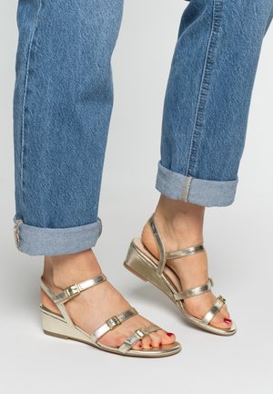 Feet wearing metallic gold wedge sandals with buckled straps, paired with rolled-up blue jeans against a plain background.