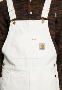 Salopette en denim blanc avec des bretelles réglables, des accents dorés et une poche frontale. Présente un patch logo Carhartt à l'avant.
