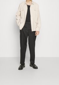 Beige colbert met kraag, zwart t-shirt eronder, losse zwarte broek, zwarte loafers met kettingdetail, minimalistisch ontwerp.