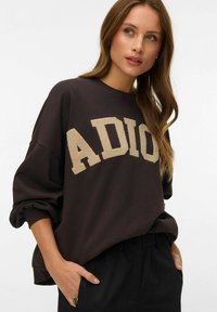 Junge Frau mit langem Haar, die einen dunklen, übergroßen Pullover mit beigen, strukturierten "ADIO"-Buchstaben trägt, die Hände in den Taschen, schaut seitlich.