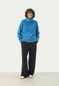 Modra turtleneck pulover z rebrastim robom in manšetami, v kombinaciji z ohlapnimi temno modrimi hlačami in belimi supergami. Gladka tekstura.