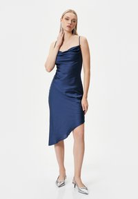 Navy Satin-Kleid mit Cowl-Ausschnitt, asymmetrischem Saum und dünnen Trägern; kombiniert mit silbernen spitz zulaufenden Absätzen und langen Ohrringen.