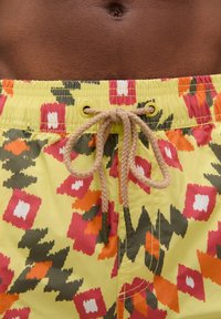 Shorts de bain jaunes à motif géométrique en rouge, orange et vert. Ils disposent d'une taille à cordon de serrage et d'une texture lisse.
