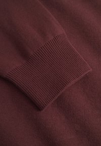 Nærbillede af en ribbet manchet på en burgunderfarvet strikket sweaterærme, der viser tekstur og stofdetaljer.