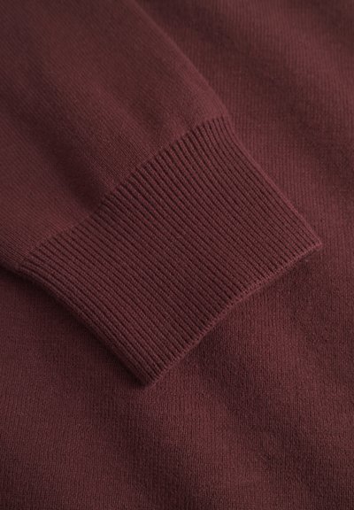 Gros plan sur une manche de pull en tricot bordeaux avec un poignet côtelé, révélant la texture et les détails du tissu.