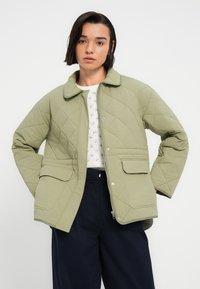 Ανοιχτό πράσινο καπιτονέ μπουφάν με γιακά, κλείσιμο με κουμπιά στο μπροστινό μέρος και δύο μεγάλες τσέπες. Διαθέτει ομαλή υφή και μοτίβο σε σχήμα διαμαντιών.