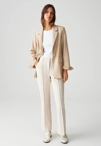 Beige blazer met een relaxed fit en notch lapels, gecombineerd met crème op maat gemaakte broeken. Witte t-shirt eronder en lichte loafers.