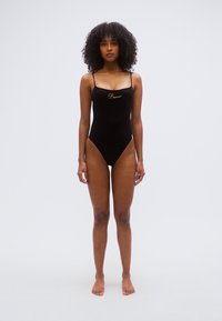 Maillot de bain une pièce noir avec bretelles fines, présentant un logo doré sur la poitrine, une texture lisse et des ouvertures hautes aux jambes.