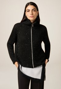 Schwarzer, gestrickter Reißverschluss-Pullover mit hohem Kragen, der ein strukturiertes Design und subtile metallische Fäden aufweist. Kombiniert mit einer grauen Bluse mit Spitzenbesatz am Saum.