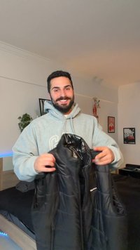 Hombre con sudadera gris con capucha sonriendo mientras sostiene un chaleco negro acolchado dentro de una habitación con cuadros enmarcados y plantas.