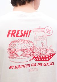 Άτομο που φορά λευκό πουκάμισο με κόκκινο γραφικό μπιφτεκιού, πατάτες σε καλάθι, ποτήρι με ετικέτα Levi's και κείμενο "ΦΡΕΣΚΟ! Δεν υπάρχει υποκατάστατο για τα κλασικά"