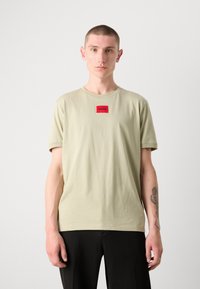 Tricou din bumbac de culoare verde deschis, cu guler rotund, mâneci scurte și o etichetă pătrată roșie cu inscripția "HUGO" pe piept.