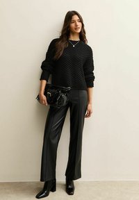 Zwarte textuurtrui met lange mouwen, gecombineerd met glanzende zwarte faux leren broeken. Model houdt een zwarte tas vast en draagt elegante zwarte enkellaarsjes.