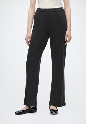 Femme portant un pantalon noir droit, un haut sans manches blanc et des ballerines en cuir verni noir, debout devant un fond blanc.