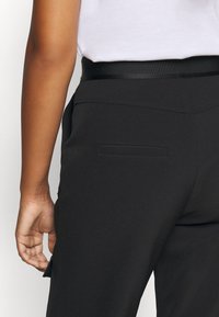 Personne portant un pantalon noir et un haut blanc, montrant la poche arrière et la zone de la taille, avec une main détendue sur le côté.
