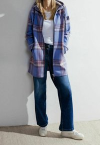 Giacca a quadri in tonalità di blu e viola con cappuccio, abbinata a jeans a gamba larga blu e sneaker bianche. Presenta tasche laterali.