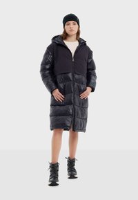 Abrigo acolchado largo negro con cremallera en el frente, capucha y secciones de tela brillante y mate. Combinado con botas negras con cordones.