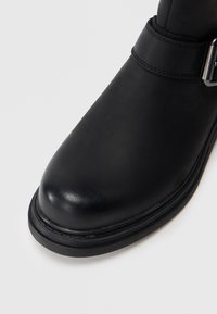 Black leather ankle boot featuring a rounded toe, matte texture, double stitching, and a strap with a silver buckle accent.