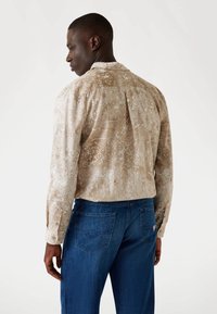 Beige, lange mouwen overhemd in een gemusterde stof met een achterste plooien. Gecombineerd met donkerblauwe spijkerbroek met twee achterzakken.