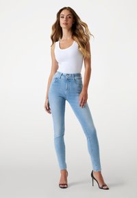 Jean skinny taille haute bleu en denim délavé avec des coutures contrastées. Associé à un débardeur blanc et des talons ouverts noirs.