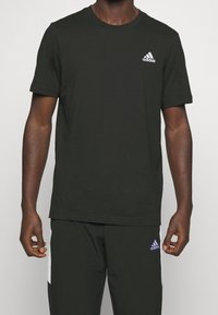 Zwarte katoenen T-shirt met een ronde hals, korte mouwen en een klein wit Adidas-logo op de linkerborst. Soepele textuur, standaard pasvorm.