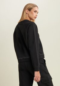 Giovane donna con capelli lunghi biondi, che indossa una felpa nera e pantaloni cargo neri, in piedi di lato contro uno sfondo semplice.