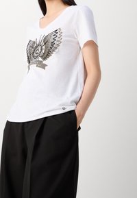 Vit kortärmad t-shirt med v-ringning, prydd med en metallisk broderad fjärilsdesign och en liten logodetalj på sidan.