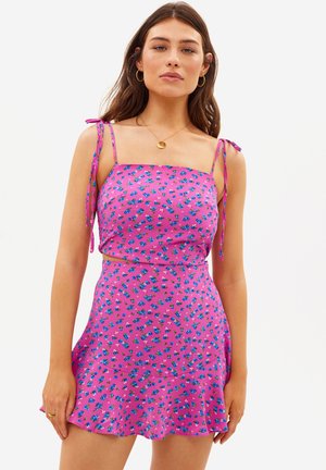 Jeune femme portant une robe sans manches rose à fleurs avec des bretelles fines à nouer et un ourlet à volants, debout devant un fond blanc uni.