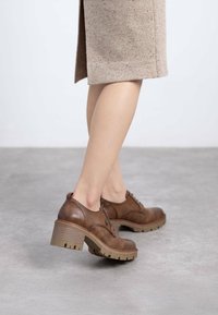 Zapatos de cuero marrón con cordones y suela de goma gruesa, con un tacón moderado, que presentan costuras en tono similar y un diseño de puntera redonda.