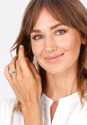 Ring mit einem zentralen grünen Edelstein, umgeben von kleinen klaren Steinen, in einem Silberschmuck gesetzt. Getragen an einer Hand mit heller Hautfarbe.