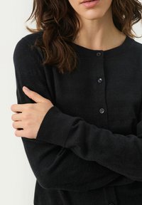 Cardigan negru din material moale, având guler rotund, închidere cu butoane și manșete cu model texturat, arătând un prim-plan cu brațele încrucișate.
