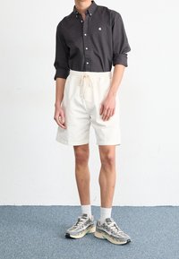 Camicia scura con bottoni e logo, pantaloncini color crema con coulisse in vita e sneakers grigie con motivo. Modello in piedi su una superficie texturizzata.