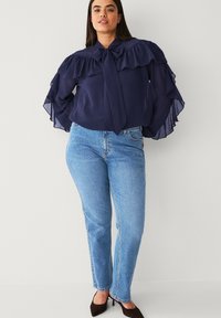 Kvinde iført en marineblå flæset bluse med sløjfe, lyseblå jeans og sorte spidse hæle, stående mod en ensfarvet lys baggrund.
