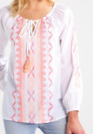 Witte blouse met lange mouwen, versierd met perzik- en roze geometrische borduursels en een voorzijde met franjestouwtje, gedragen met lichtblauwe spijkerbroek.