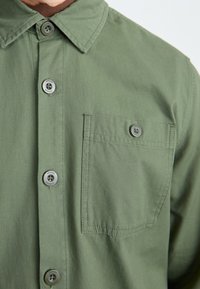 Olivenfarget knapp-up skjorte med krage, frontlomme og grå knapper. Stoffet ser jevnt ut med en subtil tekstur.