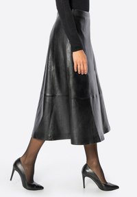 Jupe midi en cuir noir avec une coupe évasée, comprenant des détails de coutures et une texture lisse, associée à des escarpins noirs pointus et des collants transparents.