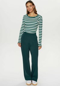 Haut à manches longues rayé en vert et blanc, associé à un pantalon large vert foncé. Modèle portant des chaussures blanches, se tenant devant un fond neutre.