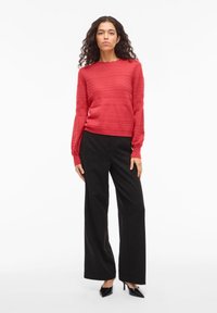 Vrouw staand, gekleed in een gestructureerde rode trui, zwarte broek met wijde pijpen en zwarte pumps met puntige neus, tegen een witte achtergrond.