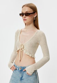 Gebreide cropped top in beige met een v-hals, lange mouwen, ruches aan de onderkant en een striksluiting. Gepaard met lichtblauwe high-waisted jeans.